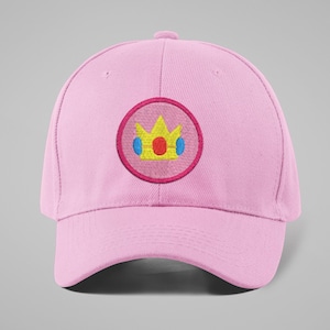 May include: A light pink baseball cap with a curved brim. The cap features a circular embroidered patch with a yellow crown design, accented with red and blue details, set against a pink background.