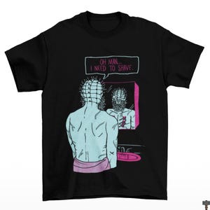 May include: Black t-shirt featuring a cartoon illustration of a character looking in a mirror. The character is depicted with a pin-covered head and is looking at a reflection of himself with a beard. Text above the mirror reads, "OH MAN... I NEED TO SHAVE."