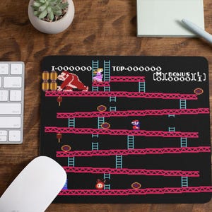 May include: Black mousepad featuring a pixelated Donkey Kong arcade game design. The game scene includes a gorilla, a princess, and Mario. The mousepad has the game's score and level displayed in white text. A white keyboard and mouse are visible.