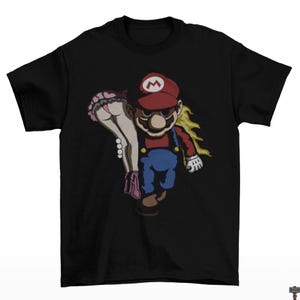 May include: Black t-shirt featuring a cartoon illustration of a character in a red hat and blue overalls carrying a woman. The woman is wearing a pink skirt and underwear. The character has a mustache and is wearing white gloves.