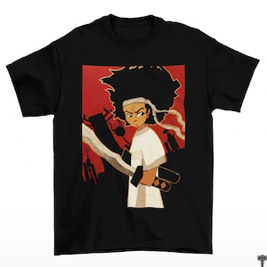 May include: Black t-shirt featuring a cartoon illustration of a character with a white headband and shirt, holding a sword. The character is set against a red backdrop with a cityscape silhouette. The design is a graphic print.