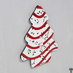 May include: A Christmas tree-shaped light switch cover. The cover is painted white with red stripes and green speckles. The light switch is in the center of the tree. The edges of the cover are black.