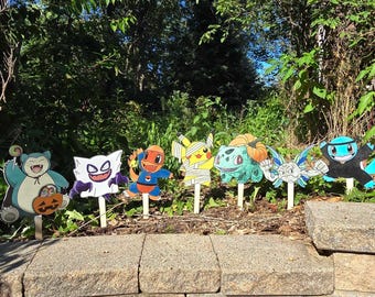 May include: A set of seven colorful character yard stakes featuring Snorlax with a pumpkin, Gengar, Charmander, Pikachu wrapped in bandages, Bulbasaur with a pumpkin, a winged creature, and a ninja-style figure, all mounted on wooden sticks.
