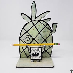 May include: A whimsical pencil holder featuring a cartoon pineapple and a character holding a yellow pencil. The pineapple is light green with black grid lines. The character is black and white, with a square body and open mouth. The pencil is yellow with a pink eraser.