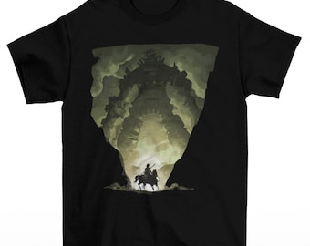 May include: Black t-shirt featuring a silhouette of a giant figure in a cave-like setting. A person on horseback is in the foreground. The graphic is in shades of green and tan, creating a dramatic contrast against the black fabric.