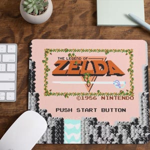 May include: A rectangular mousepad featuring the retro video game "The Legend of Zelda" design. The mousepad has a pink background with the game's logo in orange and green accents. The text "PUSH START BUTTON" is visible.