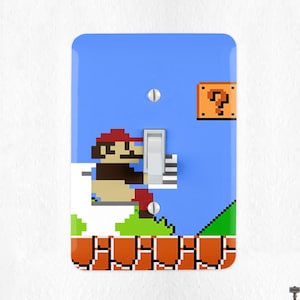 May include: A light blue wall switch cover featuring a pixelated design of a classic video game scene. The design includes a character in a red hat, a question mark block, and a brick platform. The switch is in the center.