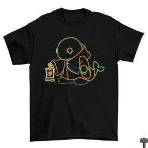 May include: Black t-shirt featuring a neon-style illustration of a hooded figure holding a lantern. The design includes green, orange, and brown outlines, with a tail-like detail. The shirt is a crew neck style.