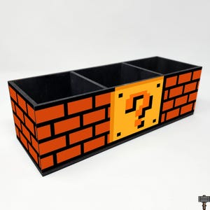May include: A rectangular wooden desk organizer with three compartments. The exterior features a pixelated design of orange bricks and a yellow question mark block, reminiscent of a classic video game. The interior is black.