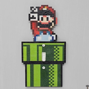 May include: A pixelated Mario character emerging from a green pipe, designed as a light switch cover. The design features a red hat, blue overalls, and a white shirt. The pipe has green and black stripes and a checkered pattern.