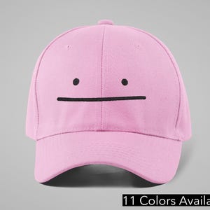 May include: A light pink baseball cap with a black embroidered face design. The cap features a curved brim and a rounded crown. The text "11 Colors Available" is displayed at the bottom of the image.