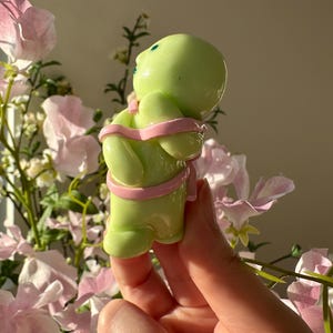 K&ouml;nnte beinhalten: Hellgr&uuml;ne Figur einer Person mit rundem Kopf und Armen, die mit einem rosa Band umwickelt sind. Die Figur hat zwei kleine blaue Punkte als Augen. Der Hintergrund zeigt rosa Blumen und Gr&uuml;n.