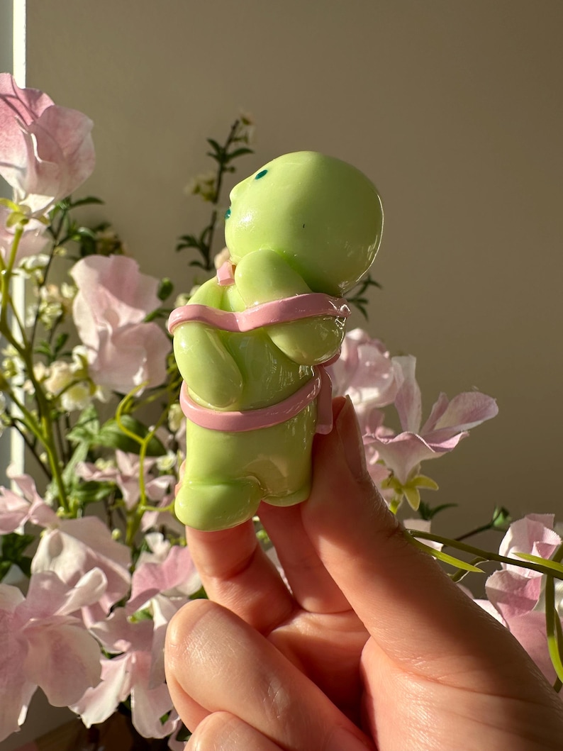 K&ouml;nnte beinhalten: Hellgr&uuml;ne Figur einer Person mit rundem Kopf und Armen, die mit einem rosa Band umwickelt sind. Die Figur hat zwei kleine blaue Punkte als Augen. Der Hintergrund zeigt rosa Blumen und Gr&uuml;n.