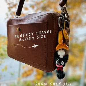 May include: A brown leather crossbody bag with a front pocket. The text "PERFECT TRAVEL BUDDY SIZE" is printed on the pocket, along with an airplane graphic. A charm of a black cat chef with a red bow tie hangs from the strap. The text "SHOW CHEF JIJI JR. THE WORLD!" is printed at the bottom.