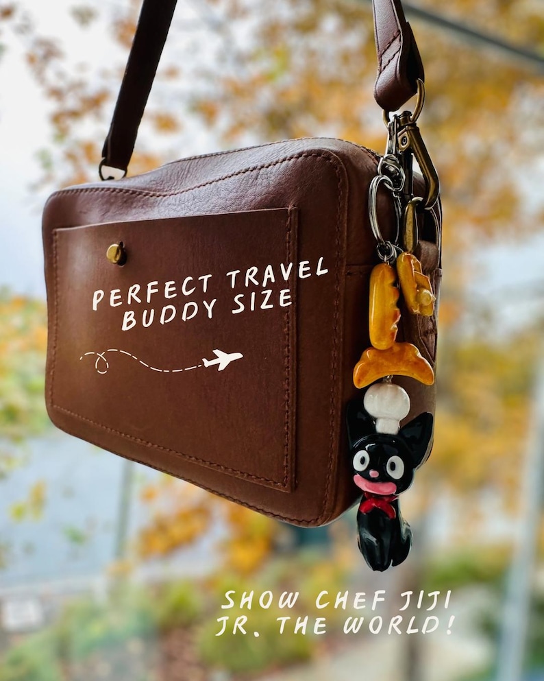 May include: A brown leather crossbody bag with a front pocket. The text "PERFECT TRAVEL BUDDY SIZE" is printed on the pocket, along with an airplane graphic. A charm of a black cat chef with a red bow tie hangs from the strap. The text "SHOW CHEF JIJI JR. THE WORLD!" is printed at the bottom.