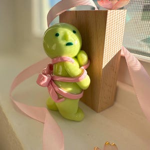 K&ouml;nnte beinhalten: Eine skurrile, gr&uuml;ne Keramikfigur mit einem rosa Band, das um sie gewickelt ist, neben einem Holzblock. Die Figur hat einen runden Kopf und kleine Arme. Ein rosa Band und eine dekorative Blume sind ebenfalls vorhanden.