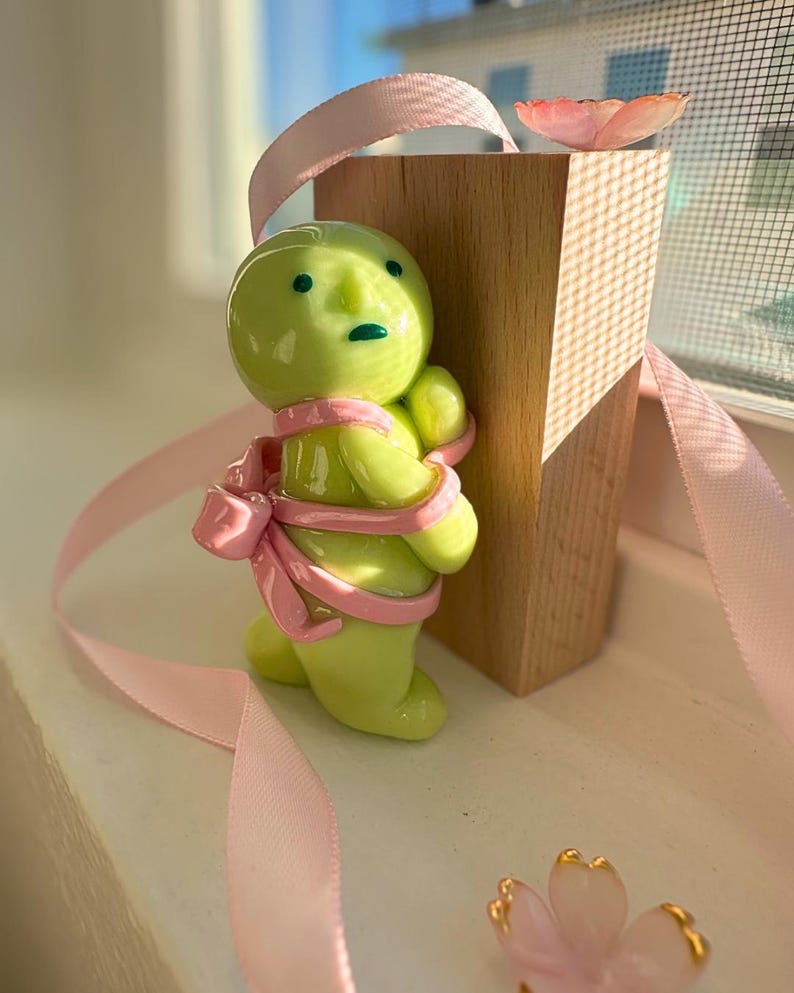 K&ouml;nnte beinhalten: Eine skurrile, gr&uuml;ne Keramikfigur mit einem rosa Band, das um sie gewickelt ist, neben einem Holzblock. Die Figur hat einen runden Kopf und kleine Arme. Ein rosa Band und eine dekorative Blume sind ebenfalls vorhanden.