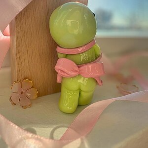 K&ouml;nnte beinhalten: Hellgr&uuml;ne Figur mit rosa Schleife und Schal. Die Figur ist neben einem Holzblock und rosa Band platziert. Eine goldene und rosa Blume befindet sich ebenfalls im Bild. Die Figur hat ein gl&auml;nzendes Finish.