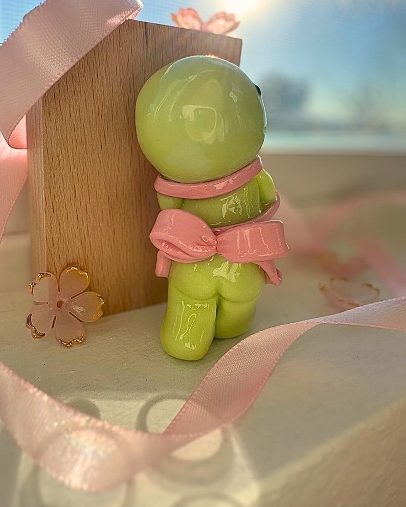 K&ouml;nnte beinhalten: Hellgr&uuml;ne Figur mit rosa Schleife und Schal. Die Figur ist neben einem Holzblock und rosa Band platziert. Eine goldene und rosa Blume befindet sich ebenfalls im Bild. Die Figur hat ein gl&auml;nzendes Finish.