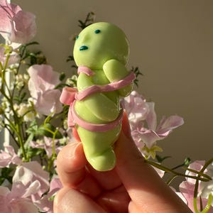 K&ouml;nnte beinhalten: Hellgr&uuml;ne Figur mit rundem K&ouml;rper, kleinen Armen und einem Kopf mit blauen Augen und Mund. Die Figur ist mit einem rosa Band und einer Schleife umwickelt. Der Hintergrund zeigt rosa und wei&szlig;e Blumen.