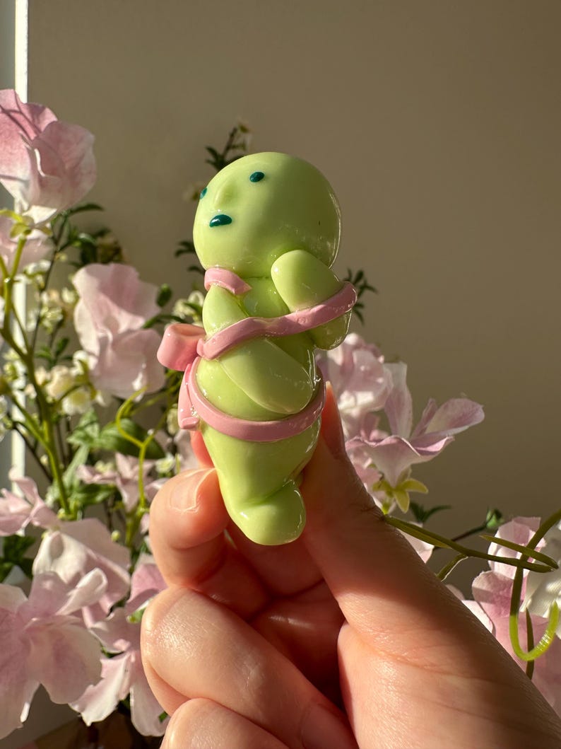 K&ouml;nnte beinhalten: Hellgr&uuml;ne Figur mit rundem K&ouml;rper, kleinen Armen und einem Kopf mit blauen Augen und Mund. Die Figur ist mit einem rosa Band und einer Schleife umwickelt. Der Hintergrund zeigt rosa und wei&szlig;e Blumen.