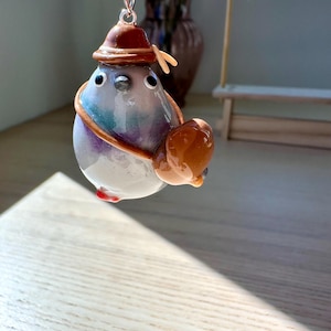 May include: A handmade keychain featuring a whimsical pigeon figurine. The pigeon is painted in shades of gray, purple, and blue, wearing a brown hat and carrying a brown bag. The keychain is attached to a silver chain.
