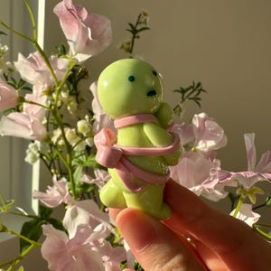 K&ouml;nnte beinhalten: Eine kleine, limettengr&uuml;ne Figur mit rundem K&ouml;rper und einer rosa Schleife darum. Die Figur hat zwei kleine, dunkle Augen und einen kleinen Mund. Der Hintergrund zeigt rosa Blumen und Gr&uuml;n.