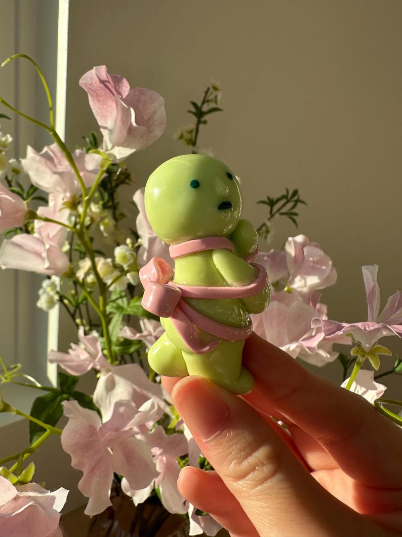 K&ouml;nnte beinhalten: Eine kleine, limettengr&uuml;ne Figur mit rundem K&ouml;rper und einer rosa Schleife darum. Die Figur hat zwei kleine, dunkle Augen und einen kleinen Mund. Der Hintergrund zeigt rosa Blumen und Gr&uuml;n.