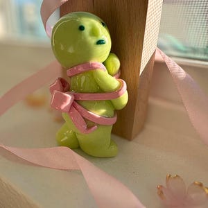 K&ouml;nnte beinhalten: Eine skurrile, lindgr&uuml;ne Figur mit einer rosa Schleife und einem Band, die einen Holzblock umarmt. Die Figur hat einen runden Kopf und kleine, dunkle Augen. Ein rosa Band ist um den K&ouml;rper der Figur gewickelt. Eine rosa Blume befindet sich oben auf dem Holzblock.