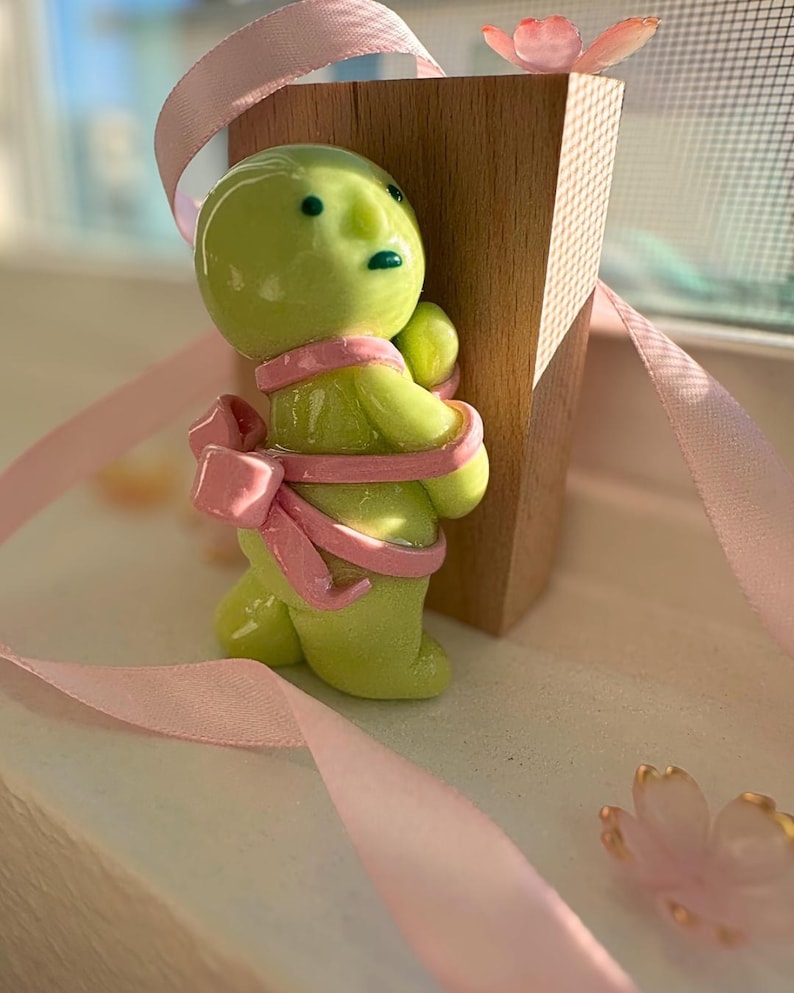 K&ouml;nnte beinhalten: Eine skurrile, lindgr&uuml;ne Figur mit einer rosa Schleife und einem Band, die einen Holzblock umarmt. Die Figur hat einen runden Kopf und kleine, dunkle Augen. Ein rosa Band ist um den K&ouml;rper der Figur gewickelt. Eine rosa Blume befindet sich oben auf dem Holzblock.