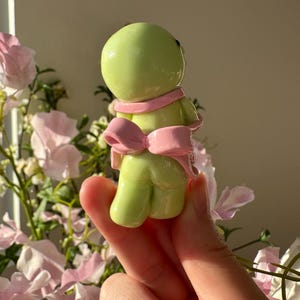 K&ouml;nnte beinhalten: Kleine, hellgr&uuml;ne Figur mit rundem Kopf und K&ouml;rper, die einen rosa Schal und eine Schleife tr&auml;gt. Die Figur wird in einer Hand gehalten, mit einem verschwommenen floralen Hintergrund aus rosa und wei&szlig;en Blumen.