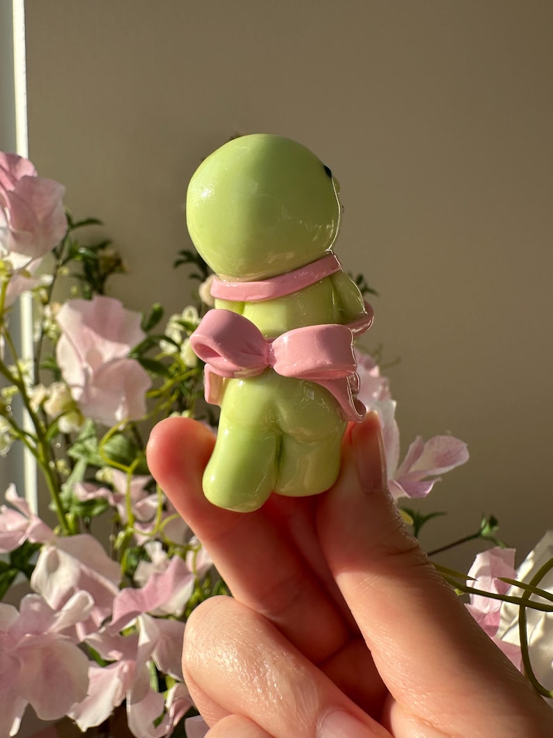 K&ouml;nnte beinhalten: Kleine, hellgr&uuml;ne Figur mit rundem Kopf und K&ouml;rper, die einen rosa Schal und eine Schleife tr&auml;gt. Die Figur wird in einer Hand gehalten, mit einem verschwommenen floralen Hintergrund aus rosa und wei&szlig;en Blumen.