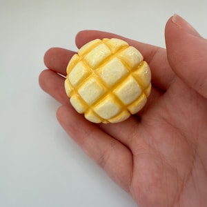 Peut inclure: Un petit objet rond jaune et crème avec un motif quadrillé, ressemblant à un melon pan. L'objet est tenu dans une main ouverte sur un fond blanc. L'objet est un aliment miniature.