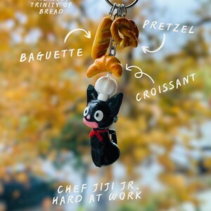 May include: A novelty keychain featuring a black cat chef holding miniature bread items: a baguette, pretzel, and croissant. The text reads "THE HOLY TRINITY OF BREAD" and "CHEF JIJI JR. HARD AT WORK."