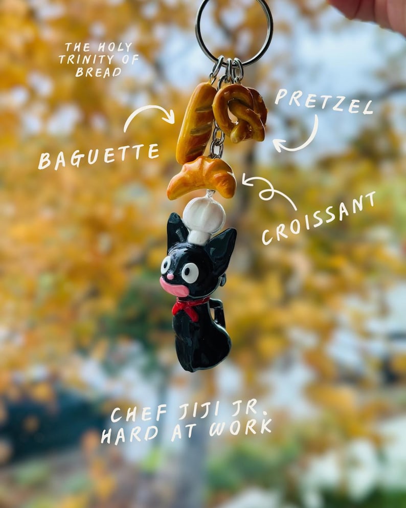 May include: A novelty keychain featuring a black cat chef holding miniature bread items: a baguette, pretzel, and croissant. The text reads "THE HOLY TRINITY OF BREAD" and "CHEF JIJI JR. HARD AT WORK."