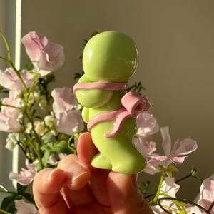 K&ouml;nnte beinhalten: Eine hellgr&uuml;ne Figur einer Person mit rundem Kopf und K&ouml;rper, umwickelt mit einem rosa Band und einer Schleife. Die Figur wird vor einem weichen Hintergrund aus rosa Blumen und Sonnenlicht gehalten.