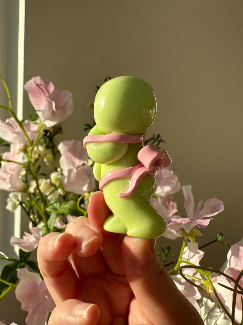 K&ouml;nnte beinhalten: Eine hellgr&uuml;ne Figur einer Person mit rundem Kopf und K&ouml;rper, umwickelt mit einem rosa Band und einer Schleife. Die Figur wird vor einem weichen Hintergrund aus rosa Blumen und Sonnenlicht gehalten.