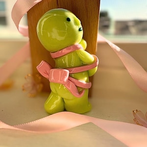 K&ouml;nnte beinhalten: Eine lindgr&uuml;ne Figur einer Person mit einer rosa Schleife um den K&ouml;rper. Die Figur hat schwarze Augen und steht vor einem Holzblock. Rosa Band und kleine rosa Bl&uuml;ten im Hintergrund.