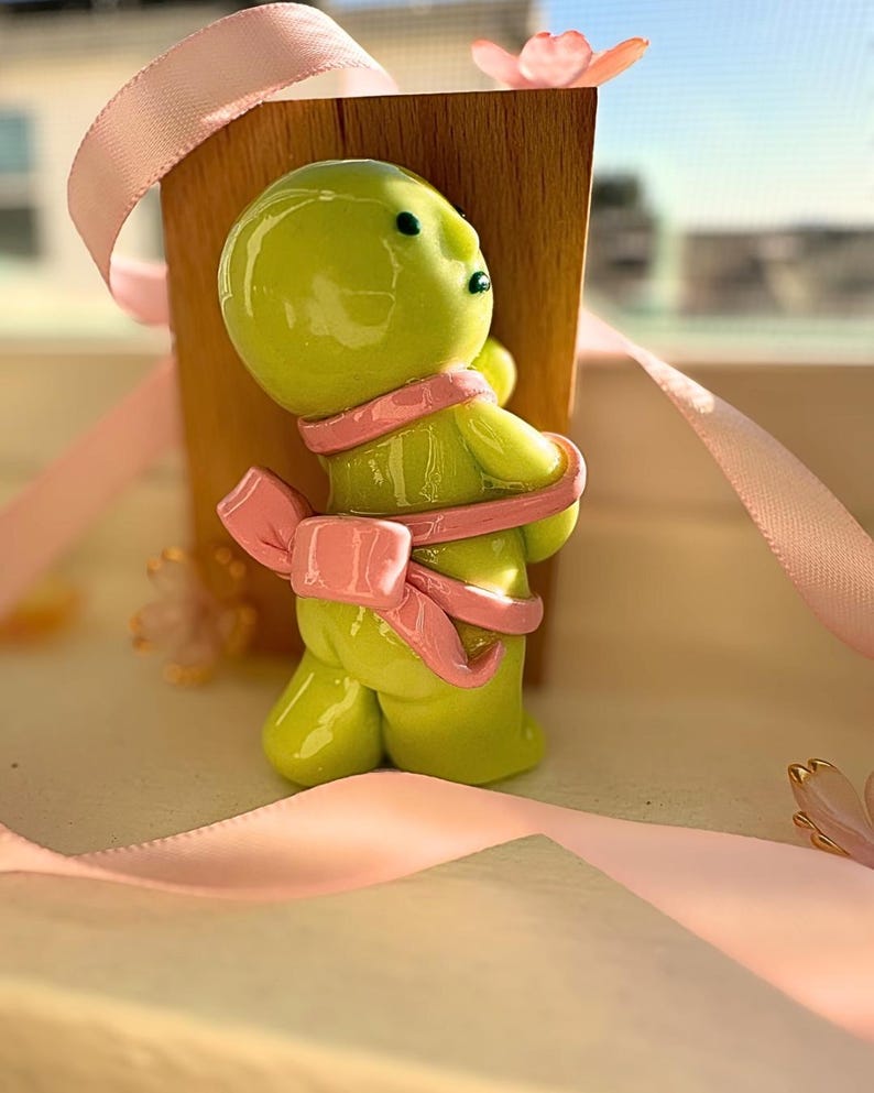 K&ouml;nnte beinhalten: Eine lindgr&uuml;ne Figur einer Person mit einer rosa Schleife um den K&ouml;rper. Die Figur hat schwarze Augen und steht vor einem Holzblock. Rosa Band und kleine rosa Bl&uuml;ten im Hintergrund.