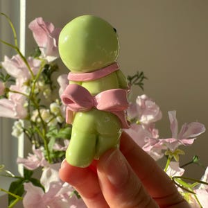 K&ouml;nnte beinhalten: Hellgr&uuml;ne Figur mit rundem Kopf und K&ouml;rper, die einen rosa Schal und eine Schleife tr&auml;gt. Die Figur wird in einer Hand gehalten, mit einem verschwommenen floralen Hintergrund aus rosa und wei&szlig;en Blumen.