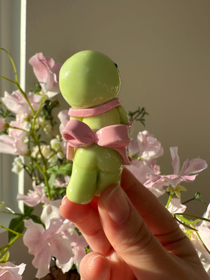 K&ouml;nnte beinhalten: Hellgr&uuml;ne Figur mit rundem Kopf und K&ouml;rper, die einen rosa Schal und eine Schleife tr&auml;gt. Die Figur wird in einer Hand gehalten, mit einem verschwommenen floralen Hintergrund aus rosa und wei&szlig;en Blumen.