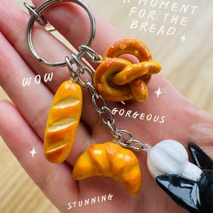 May include: A silver keyring featuring miniature food charms. The charms are a baguette, pretzel, and croissant, in shades of yellow and brown. The image includes the text "A moment for the bread", "Wow", "Gorgeous", and "Stunning".