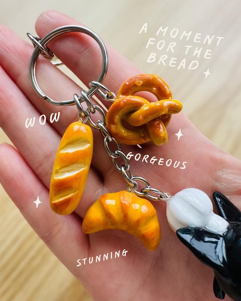 May include: A silver keyring featuring miniature food charms. The charms are a baguette, pretzel, and croissant, in shades of yellow and brown. The image includes the text "A moment for the bread", "Wow", "Gorgeous", and "Stunning".