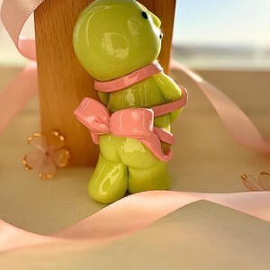 K&ouml;nnte beinhalten: Eine skurrile, limettengr&uuml;ne Figur einer Kreatur mit rosa Schal und Schleife. Die Figur ist vor einem Holzblock platziert und mit rosa B&auml;ndern und floralen Akzenten verziert, was eine verspielte und dekorative &Auml;sthetik erzeugt.