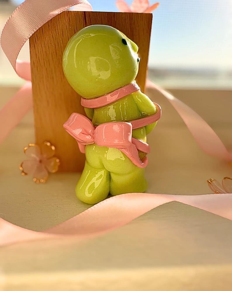 K&ouml;nnte beinhalten: Eine skurrile, limettengr&uuml;ne Figur einer Kreatur mit rosa Schal und Schleife. Die Figur ist vor einem Holzblock platziert und mit rosa B&auml;ndern und floralen Akzenten verziert, was eine verspielte und dekorative &Auml;sthetik erzeugt.