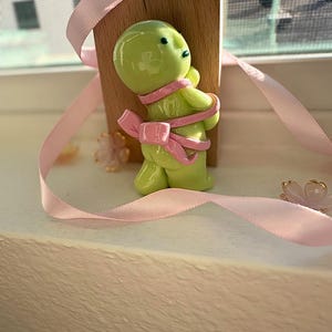 K&ouml;nnte beinhalten: Eine hellgr&uuml;ne Figur, die mit einem rosa Band mit einer Schleife auf dem R&uuml;cken umwickelt ist. Die Figur steht vor einem kleinen Holzblock. Rosa B&auml;nder und kleine Blumendekorationen sind um die Figur herum verstreut.