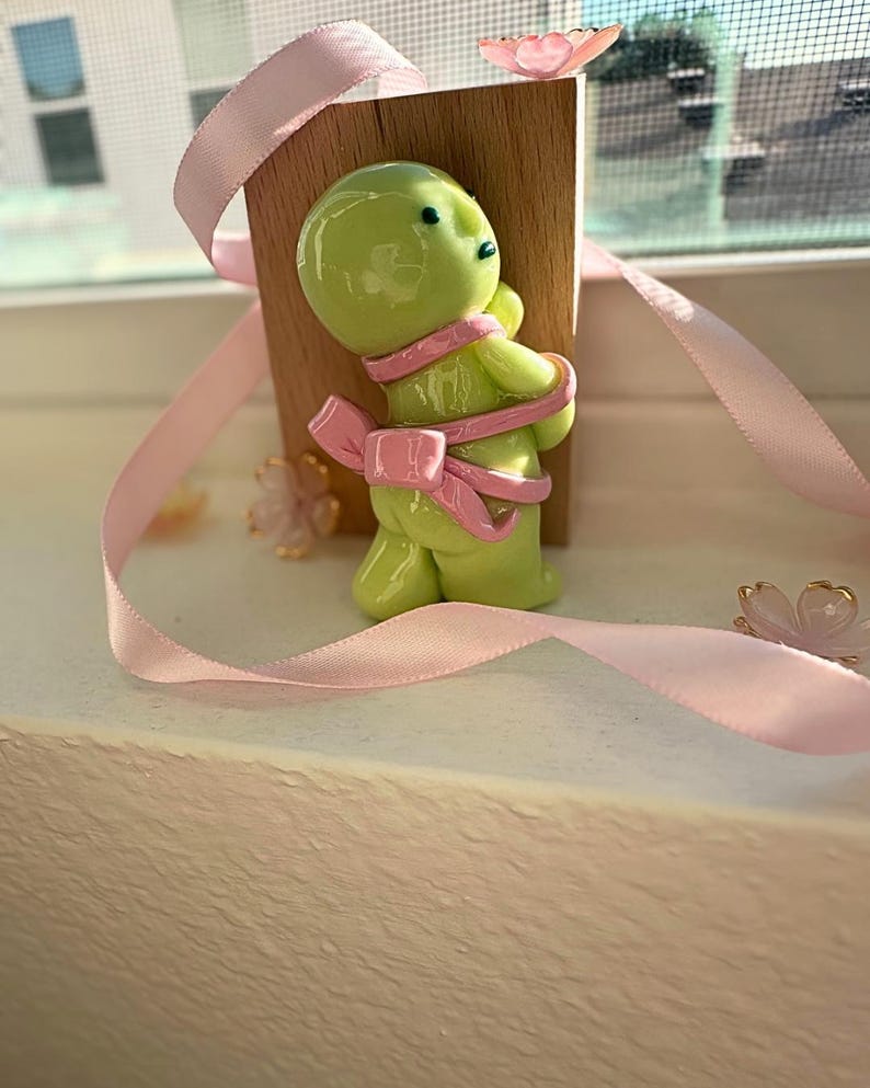 K&ouml;nnte beinhalten: Eine hellgr&uuml;ne Figur, die mit einem rosa Band mit einer Schleife auf dem R&uuml;cken umwickelt ist. Die Figur steht vor einem kleinen Holzblock. Rosa B&auml;nder und kleine Blumendekorationen sind um die Figur herum verstreut.