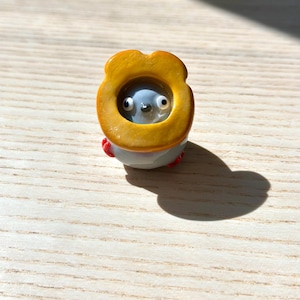 May include: A small, whimsical figurine of a creature with a gray body, large eyes, and a yellow ring around its head. The figurine has small red accents and is set against a light wood grain surface.
