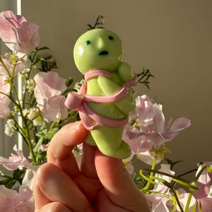 K&ouml;nnte beinhalten: Eine hellgr&uuml;ne Figur mit rundem Kopf, kleinen blauen Augen und einem rosa Band um den K&ouml;rper. Die Figur wird vor rosa Blumen gehalten.