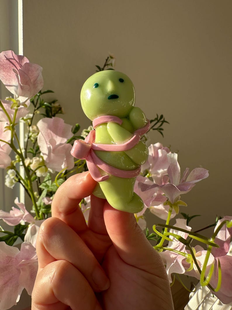 K&ouml;nnte beinhalten: Eine hellgr&uuml;ne Figur mit rundem Kopf, kleinen blauen Augen und einem rosa Band um den K&ouml;rper. Die Figur wird vor rosa Blumen gehalten.